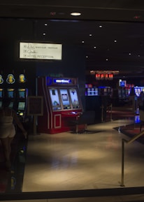 A dimly lit casino with several rows of slot machines, some displaying bright graphics and lights. There are a few people scattered throughout the area, engaging with the machines. The floor is reflective and polished, adding to the ambiance, while the ceiling is dotted with numerous small lights.