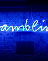 A bright blue neon sign spells out the word 'gambling' in cursive script. The background features a textured wall covered in domino tiles, also illuminated by the blue light, creating a vivid and visually striking scene.