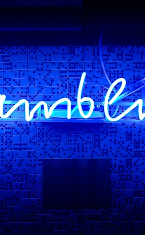A bright blue neon sign spells out the word 'gambling' in cursive script. The background features a textured wall covered in domino tiles, also illuminated by the blue light, creating a vivid and visually striking scene.