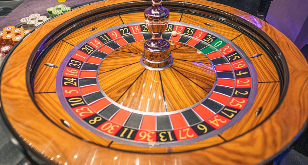 A roulette wheel with alternating red and black numbered pockets on a wooden platform. The wheel contains a small white ball resting in one of the pockets, indicating gameplay. Surrounding the wheel are stacks of colorful poker chips, suggesting a casino environment.