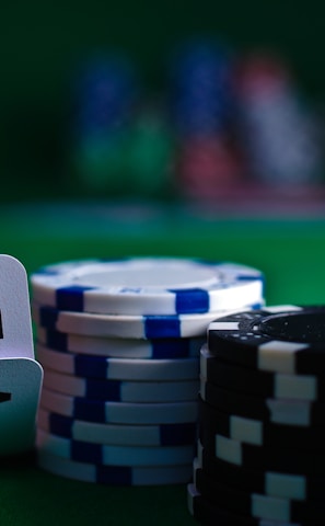 A casino scene featuring two playing cards, the ace and king of clubs, partially concealed by a player's hand. Nearby, there are stacks of multicolored poker chips on a green felt table, with blurred colorful background elements suggesting other chip stacks or cards.