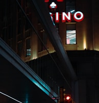 A night scene featuring a building with illuminated neon signage displaying 'CASINO'. The environment is urban with reflections on glass windows and glowing street traffic lights below. The atmosphere is dimly lit, with artificial lighting casting vibrant colors.